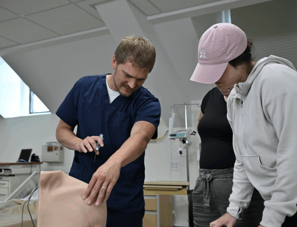 A man in medical scrubs demonstrates a procedure on a medical training mannequin arm while a person in a pink cap and hoodie observes closely in a clinical training room.
